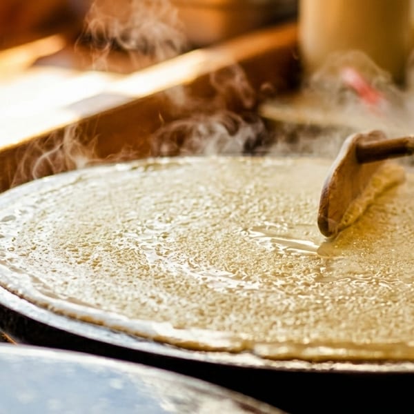A steaming crepe is spread thin on a hot griddle with a wooden T-shaped tool.