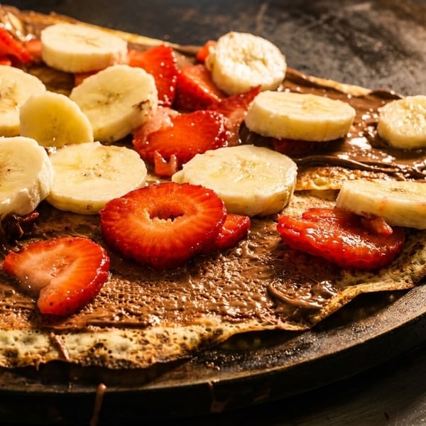 A crepe covered in chocolate spread, sliced bananas, and strawberries on a griddle.