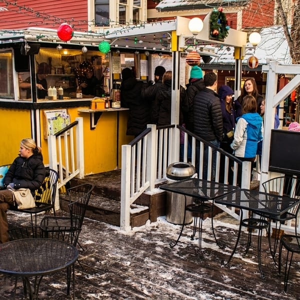 People gather around a yellow "Crêpes à la Cart" food stand with outdoor seating on a snowy day.